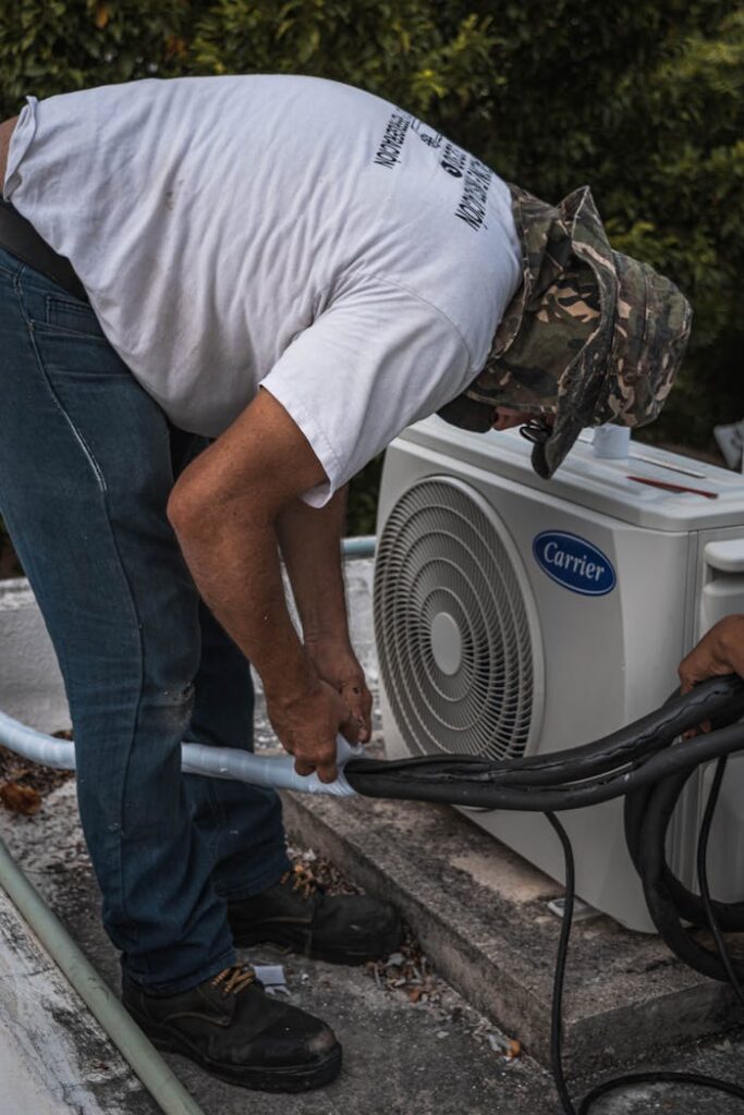 A technician performs maintenance on an outdoor air conditioning unit, focusing on hose connections.